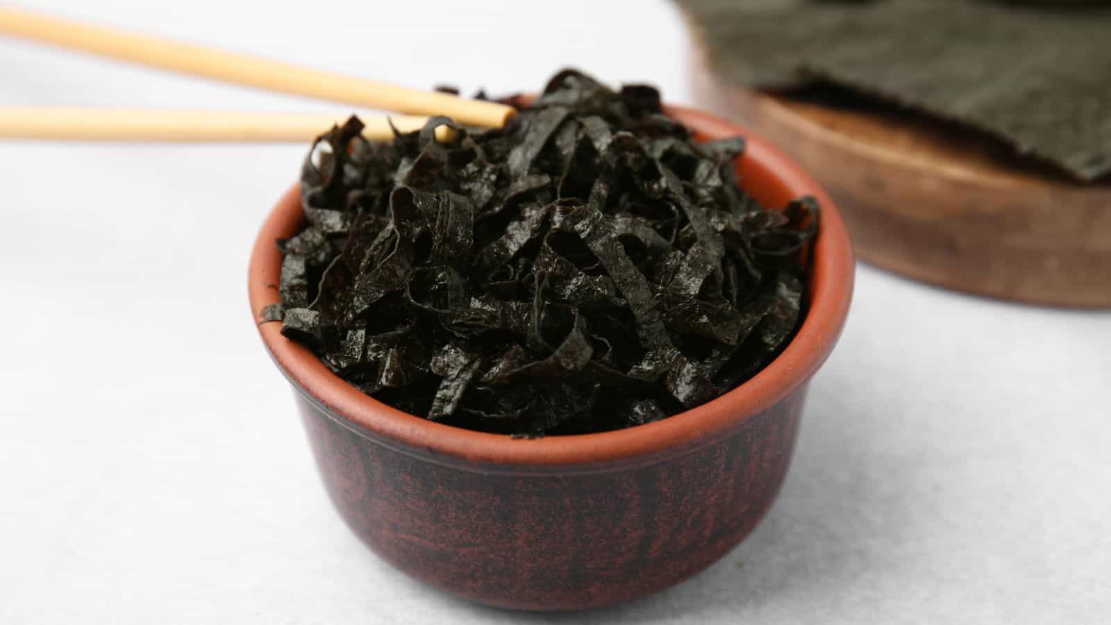 A small brown bowl filled with shredded seaweed is placed on a white surface. A pair of wooden chopsticks rests on the edge of the bowl. In the background, larger pieces of seaweed are on a wooden board.