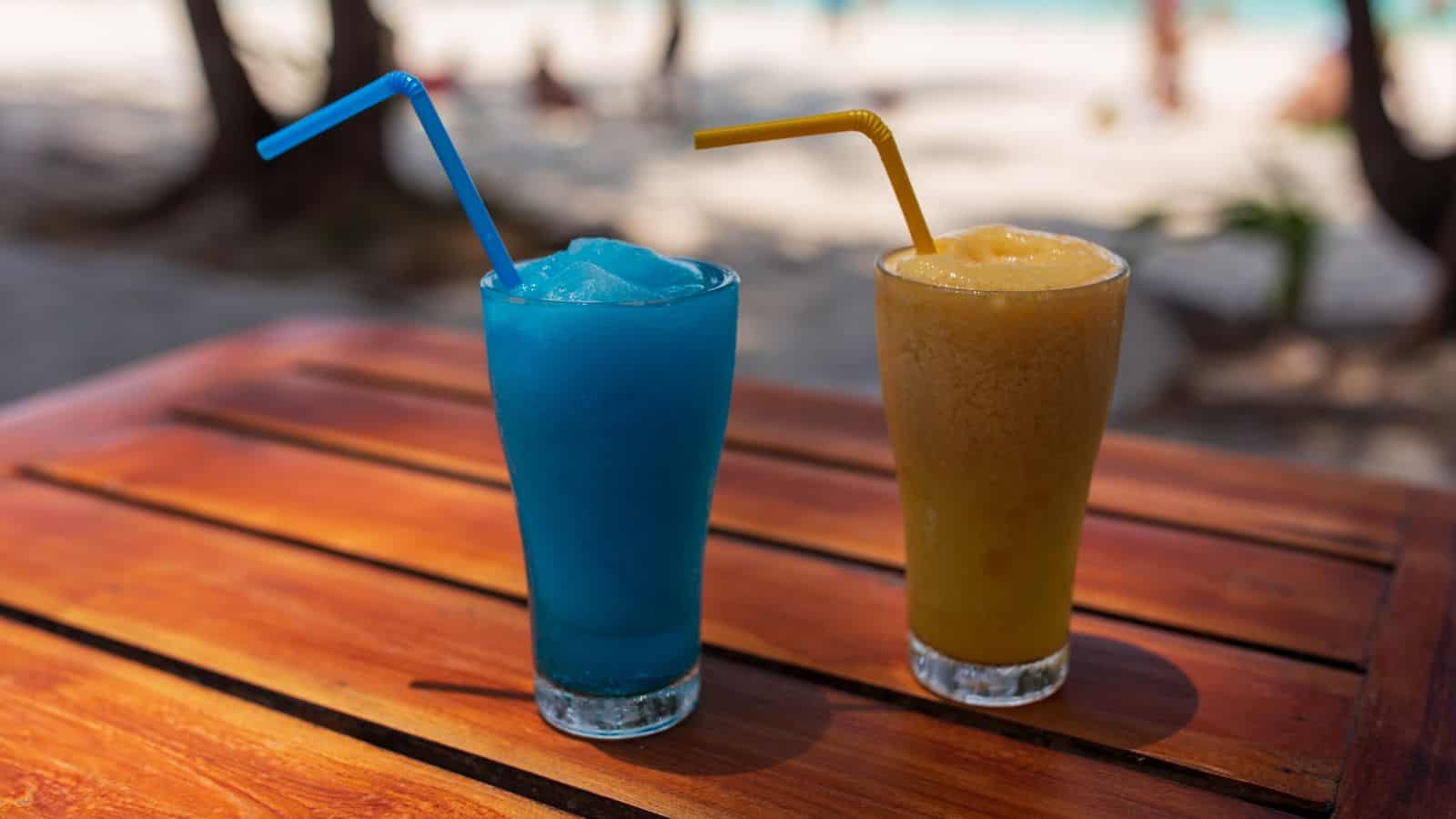 Two frosty beverages sit on a wooden table. One is a blue slushy drink with a blue straw, and the other is a yellow smoothie with a yellow straw. A beach environment is blurred in the background.