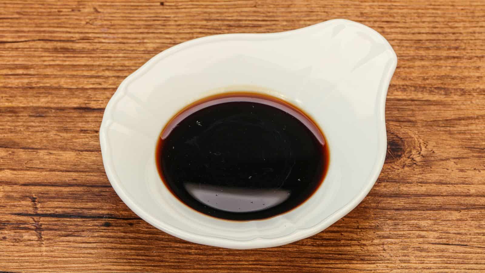 A small white ceramic dish containing dark soy sauce sits on a wooden surface. The dish has a unique, rounded shape with a slight lip, and the soy sauce fills a portion of it, displaying a glossy texture.