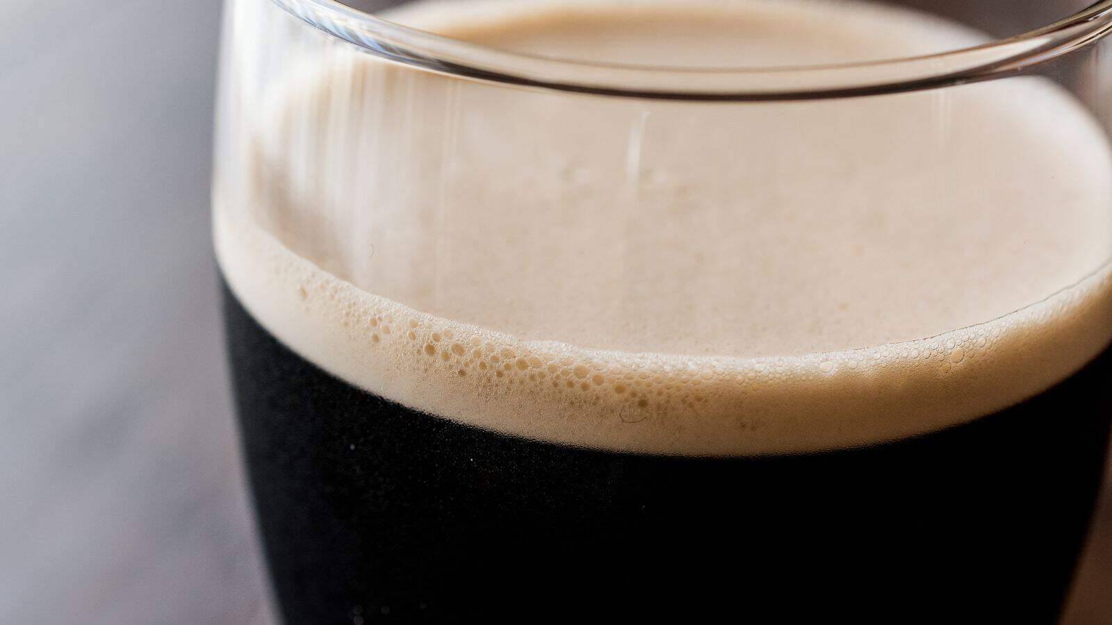 A close-up of a glass of dark beer with a creamy, frothy head. The glass is partially filled, and the beer appears rich and opaque. The background is softly blurred, emphasizing the smooth texture of the beer's surface.