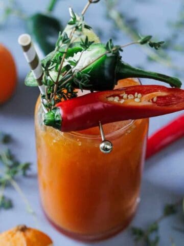 A glass filled with an orange beverage garnished with sliced chili peppers, thyme, and a straw. Surrounding the glass are whole oranges, a half lime, additional peppers, and sprigs of thyme on a light blue surface.