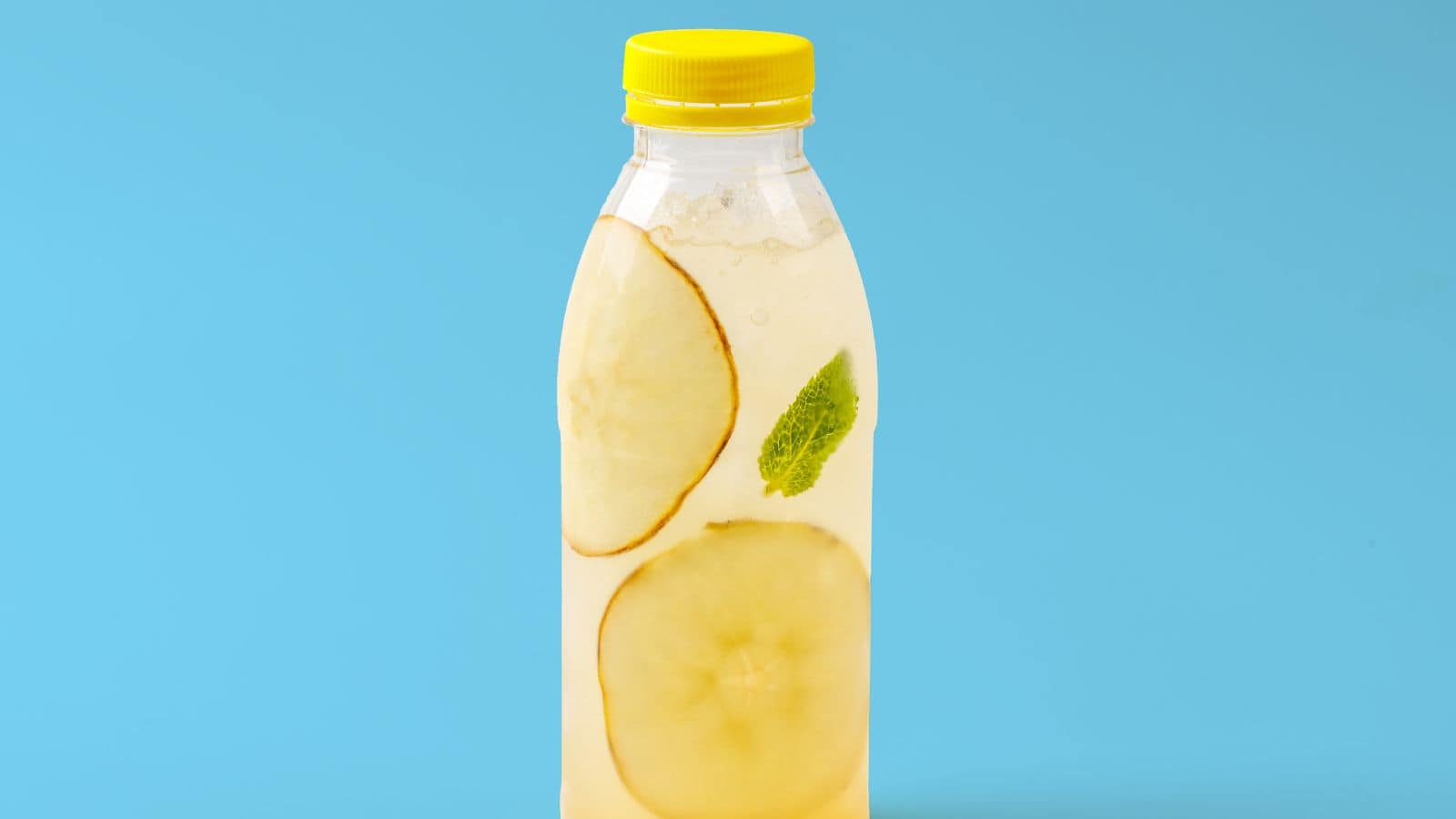 A clear bottle filled with a light yellow liquid is set against a blue background. The bottle contains slices of apple and a mint leaf, and is topped with a bright yellow cap.