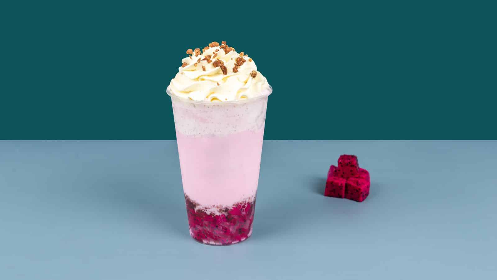 A pink beverage in a clear cup topped with whipped cream and small brown crumbs. The bottom layer contains red and pink chunks. A few red cube pieces are placed on the surface to the right. The background is teal, and the surface is light blue.