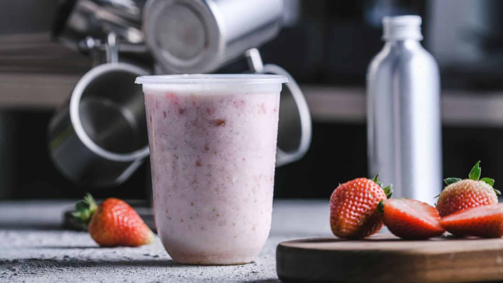 A plastic container filled with pink frozen yogurt sits on a gray surface, with a few strawberries on a wooden board in the foreground. A metal canister and cups are blurred in the background.