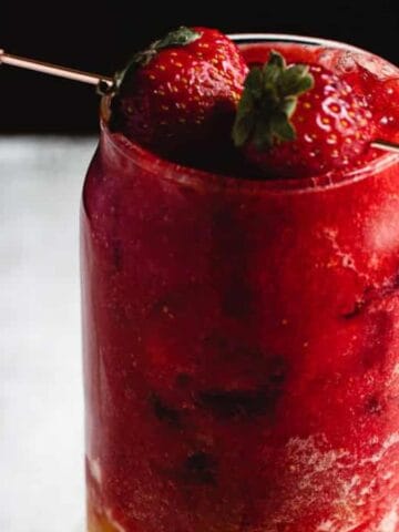 A glass filled with a red slushy drink, topped with whole strawberries on a skewer. The surface below is light and slightly textured, contrasting with the dark background.