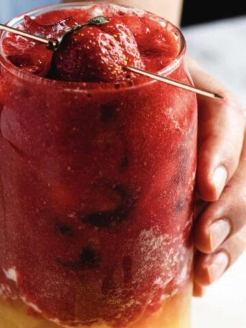 A hand holds a glass filled with a layered drink. The top layer is a thick, red berry mixture garnished with a whole strawberry and a small green leaf on a skewer. The bottom layer is a yellow liquid. The background is softly blurred.
