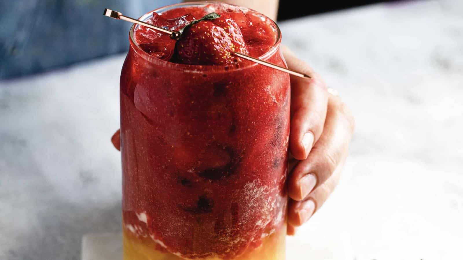 A glass filled with a red slushy drink, topped with whole strawberries on a skewer. The surface below is light and slightly textured, contrasting with the dark background.