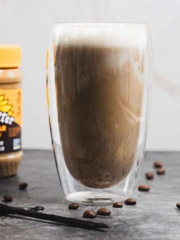 A glass of iced coffee with foam on top sits on a dark surface, surrounded by scattered coffee beans. A jar of SunButter "No Sugar Added" sunflower seed butter is in the background. Two vanilla pods are placed on the surface.