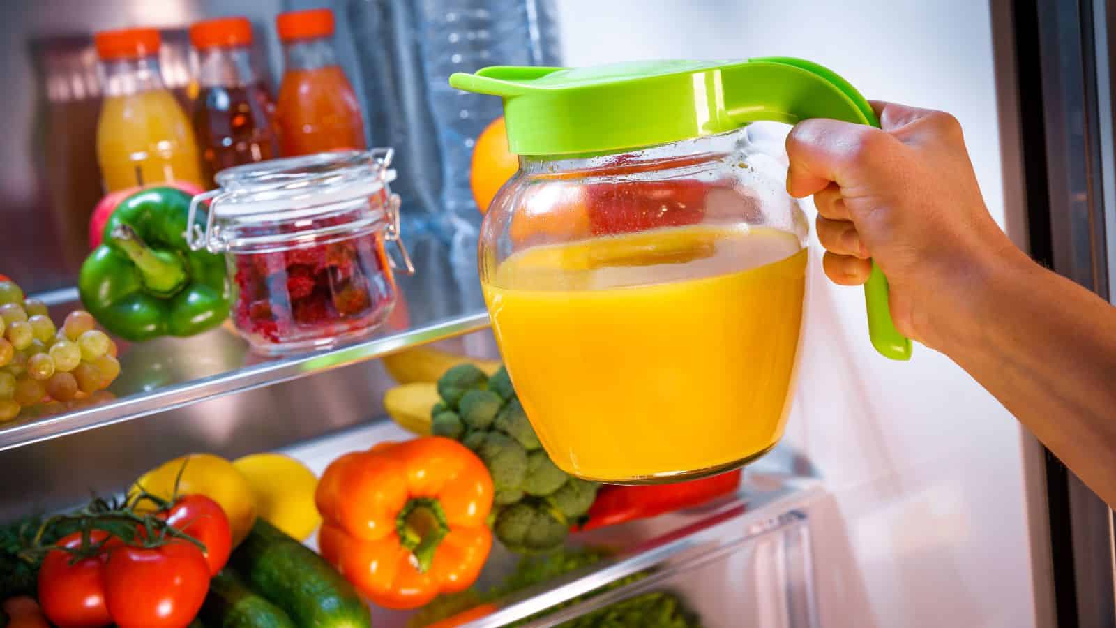 A hand holding a clear pitcher with a green lid filled with orange juice is reaching into a refrigerator. The fridge contains bottles of juice, a jar with red contents, and various fruits and vegetables like grapes, bell peppers, and broccoli.