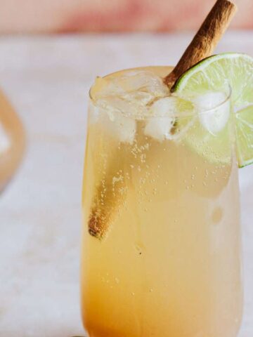 A tall glass filled with a light amber beverage, garnished with a cinnamon stick and a lime slice, sits on a light, textured surface. In the background, there is a small carafe containing a similar liquid.