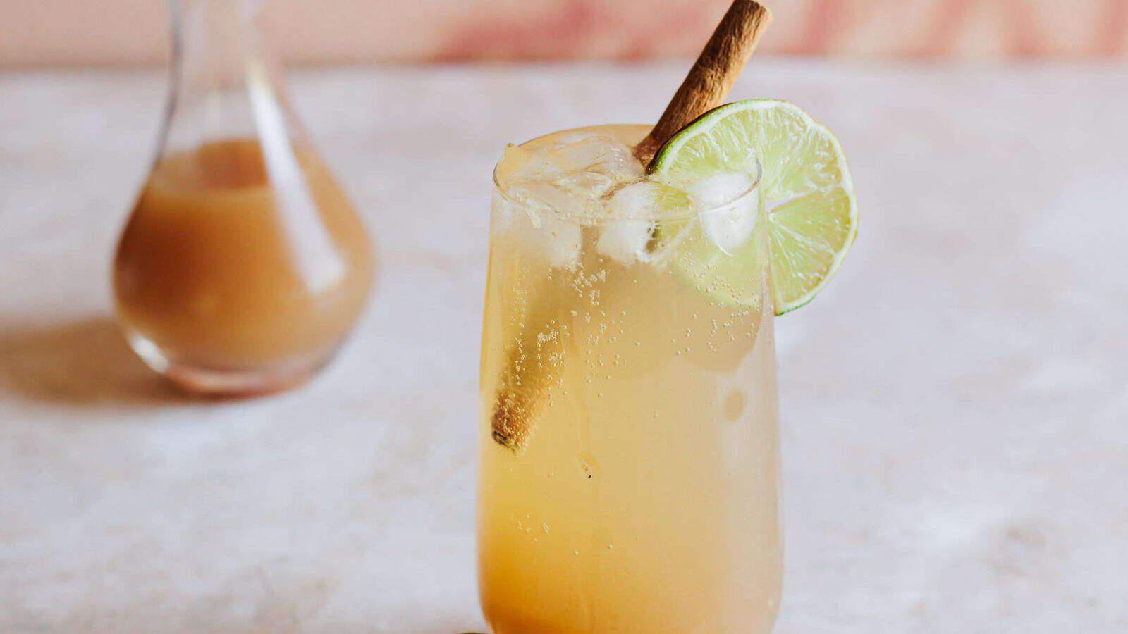 A tall glass filled with a light amber beverage, garnished with a cinnamon stick and a lime slice, sits on a light, textured surface. In the background, there is a small carafe containing a similar liquid.