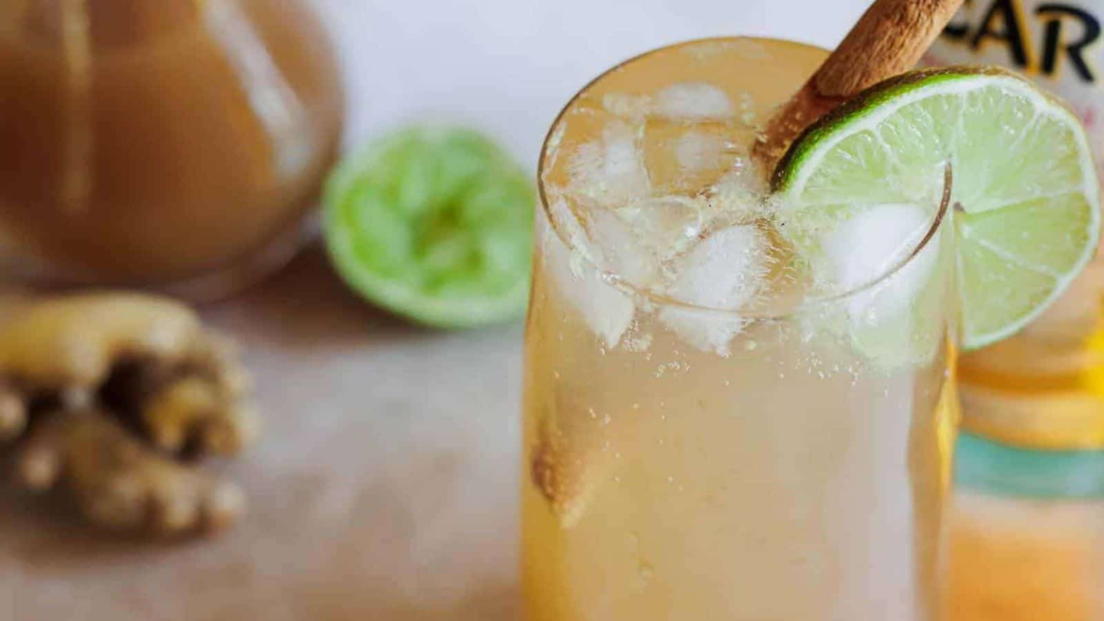 A glass filled with a light brown iced drink sits on a surface. The drink is garnished with a lime slice and a cinnamon stick. In the background, there&rsquo;s fresh ginger, half a lime, and a bottle partially visible.