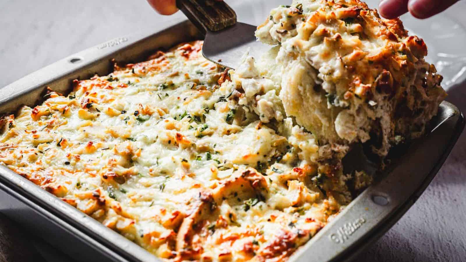 A person is using a spatula to serve a portion of baked mashed potato casserole from a metal baking dish. The casserole is topped with melted cheese and sprinkled with herbs.