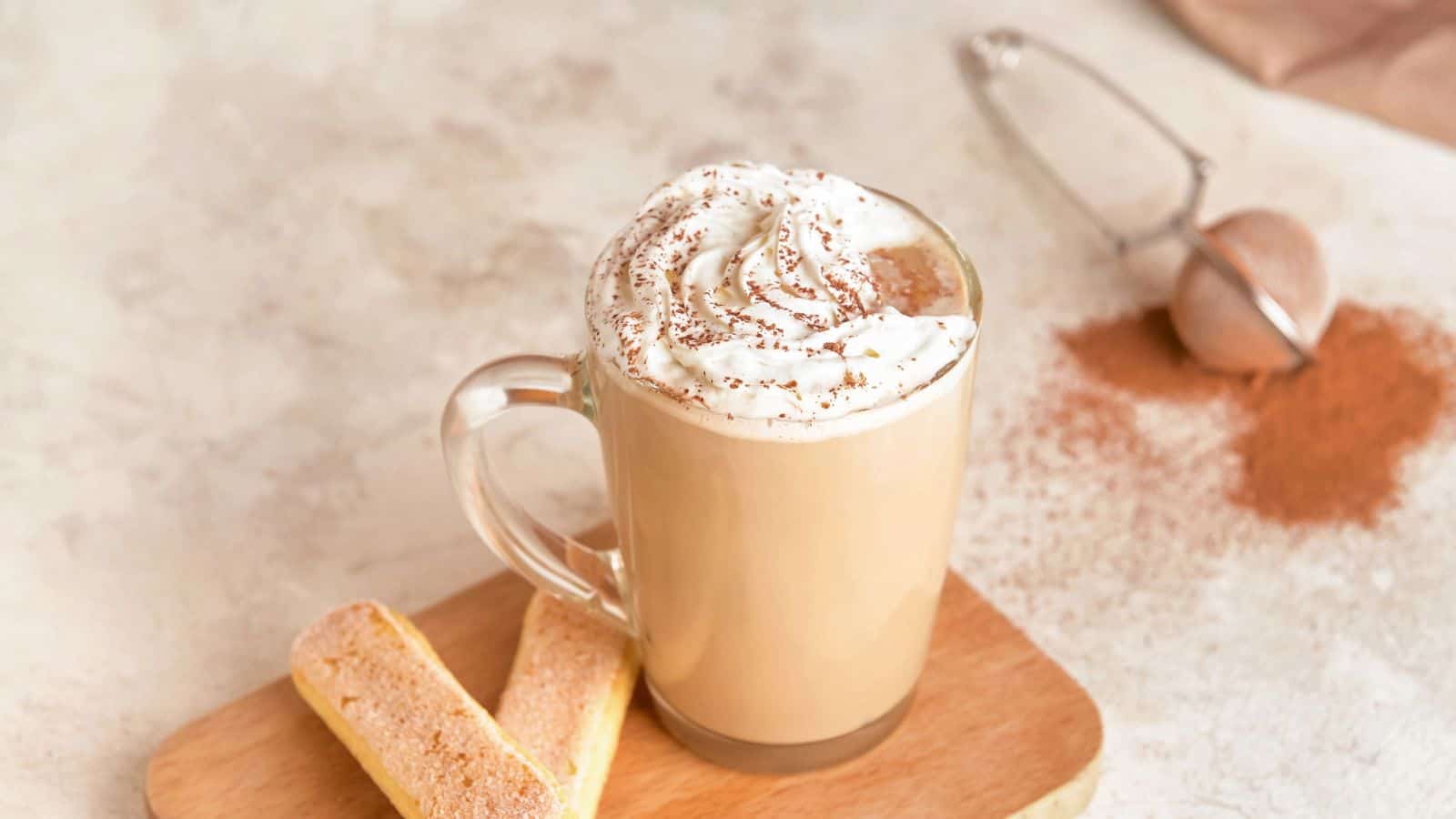 A glass mug filled with coffee topped with whipped cream and a sprinkle of cocoa sits on a wooden board. Next to it are two ladyfinger biscuits. In the background, a strainer with spilled cocoa powder is visible on a light surface.