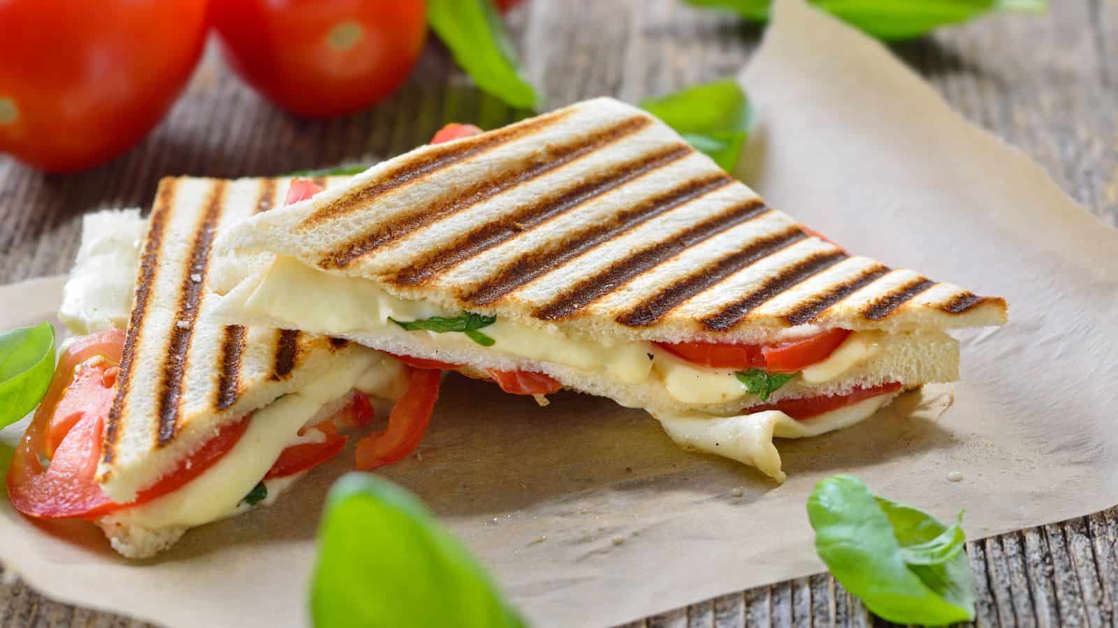 Two grilled sandwich halves on parchment paper contain melted cheese, tomato slices, and basil leaves. Grill marks are visible on the bread. In the background, there are whole tomatoes and more basil leaves scattered around.