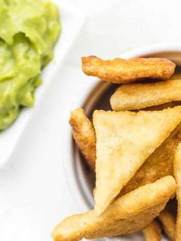 A bowl of triangular, golden-fried pita chips is placed next to a small dish of green guacamole on a white surface.
