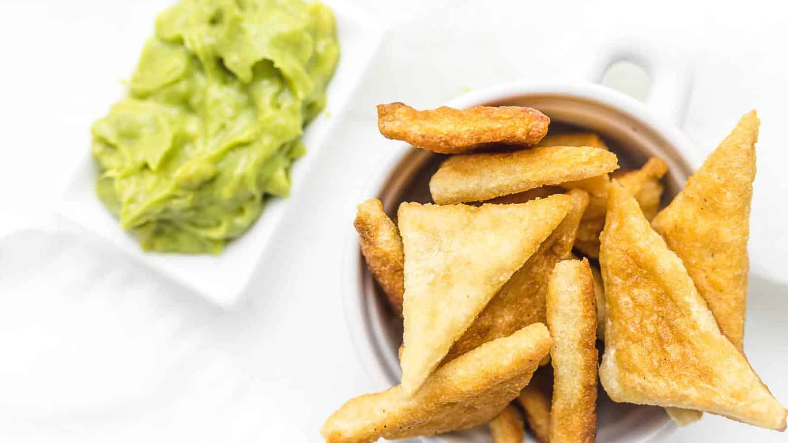 A bowl of triangular, golden-fried pita chips is placed next to a small dish of green guacamole on a white surface.