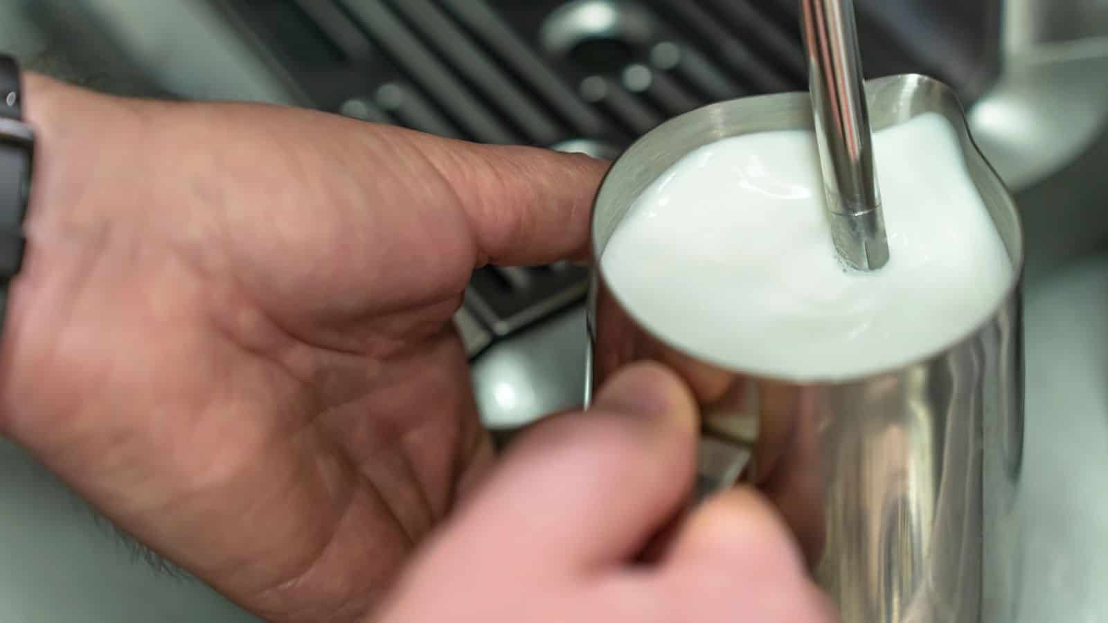 A person is frothing milk with a steam wand in a metal pitcher. The hand on the left is holding the pitcher, while the other hand adjusts the wand. The background shows a section of a coffee machine.