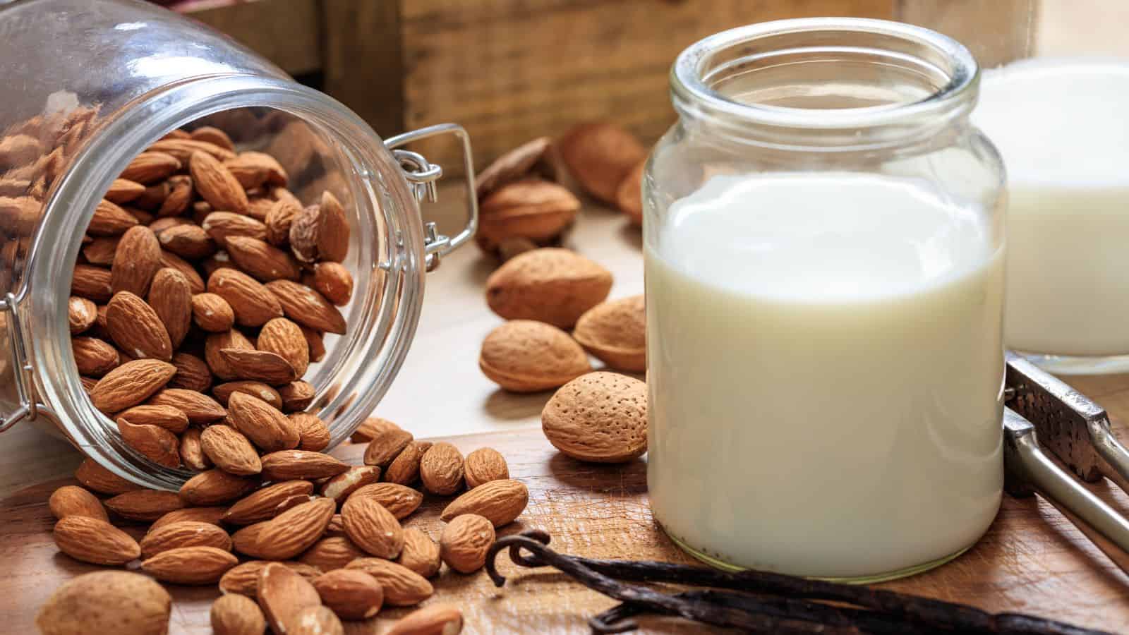 A glass jar filled with milk is placed on a wooden surface next to an open jar of almonds. Some almonds have spilled out onto the table. Vanilla pods and whole almonds with shells are also visible in the background.