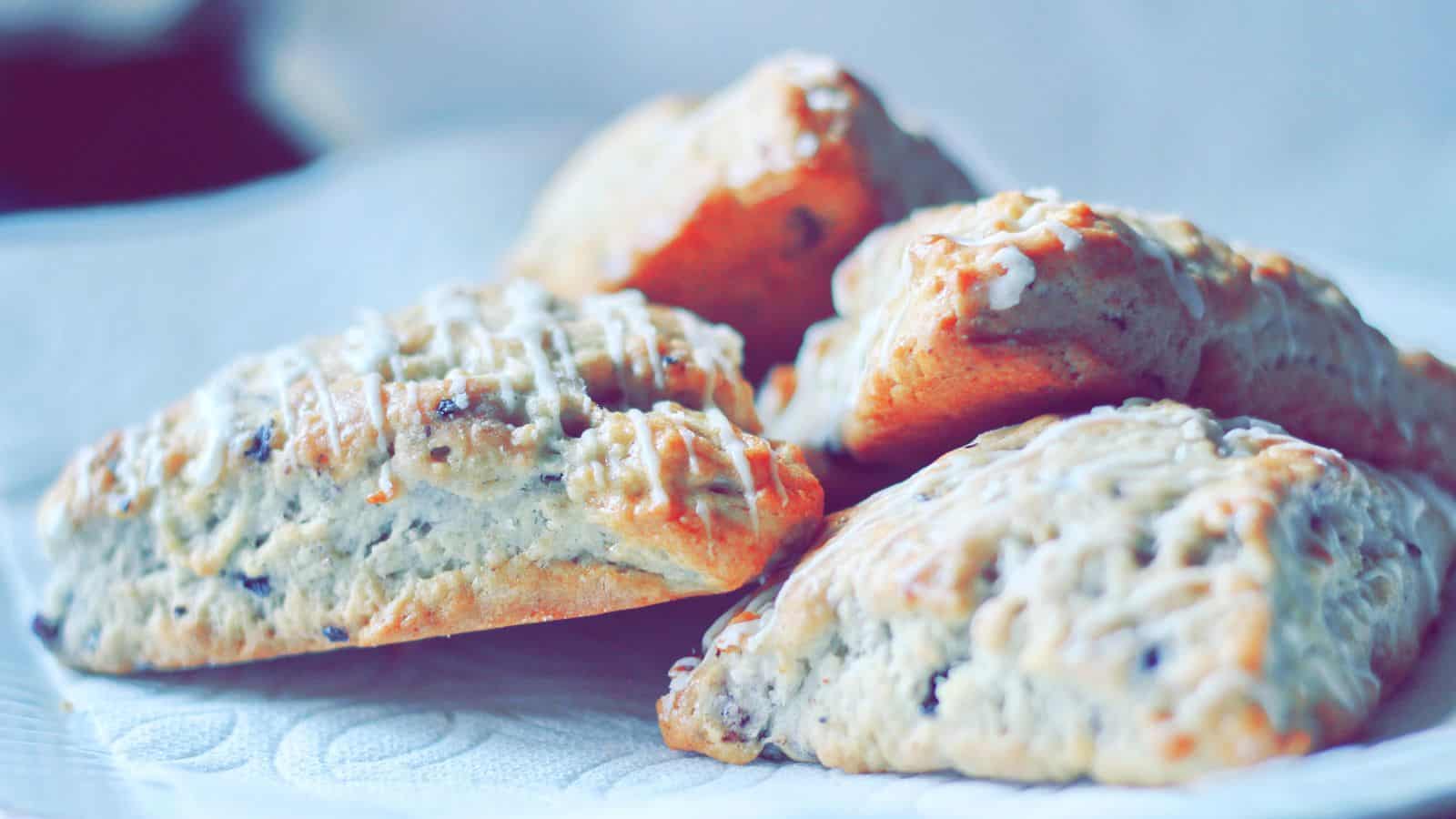 Four scones with a light drizzle of icing are placed on a white surface. The scones are golden brown with a visible texture, suggesting bits of fruit or nuts. The background is softly blurred.
