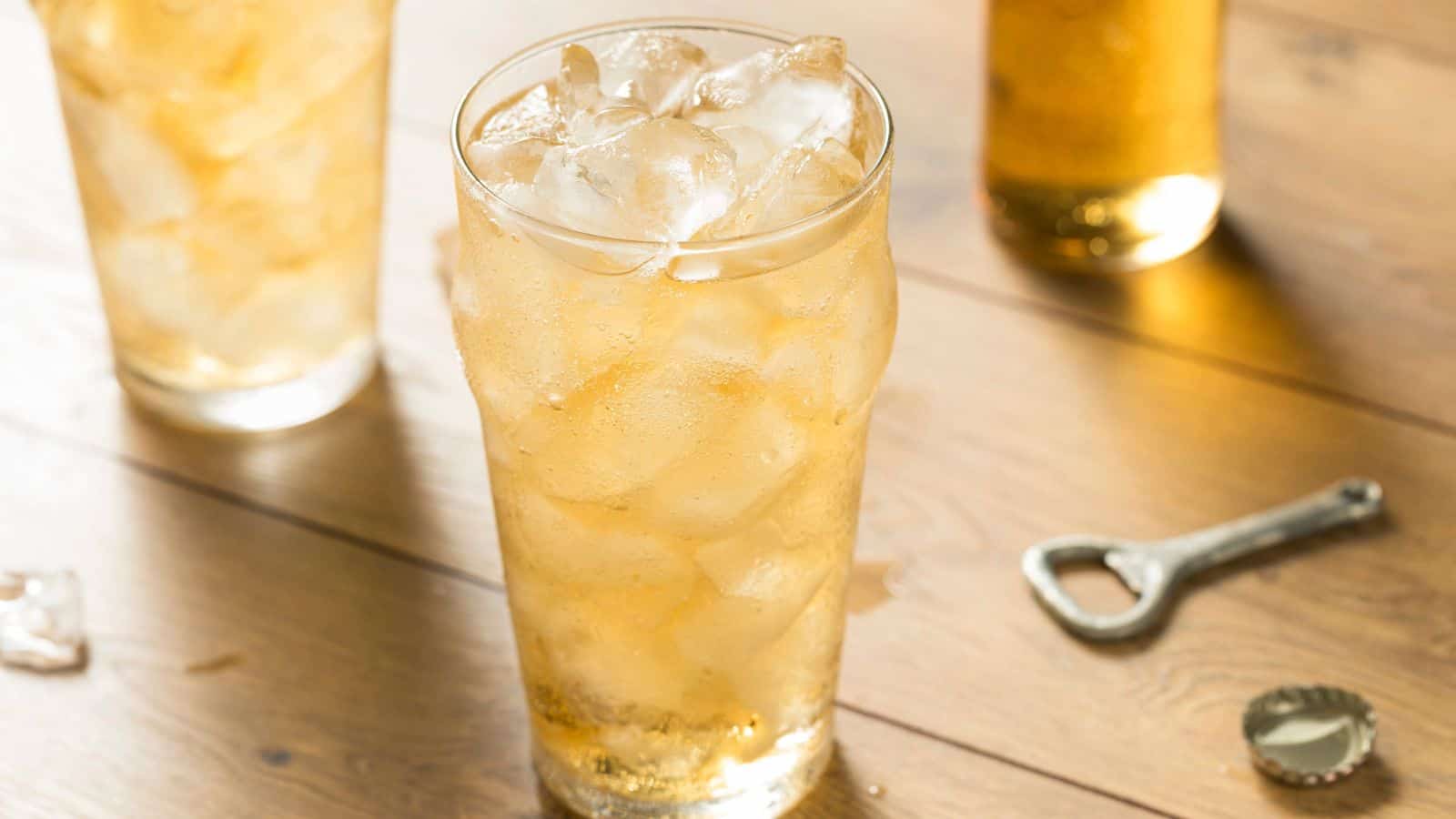 A tall glass filled with iced tea and ice cubes sits on a wooden surface. Two other glasses and a metal bottle opener with a cap are also visible. The lighting gives the scene a warm, inviting ambiance.