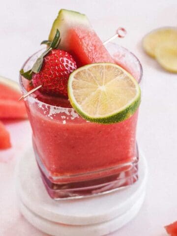 A glass of pink smoothie garnished with a watermelon slice, lime wheel, and strawberry on a skewer stands on white coasters. Surrounding it are strawberries, watermelon slices, and a lime slice on a white surface.