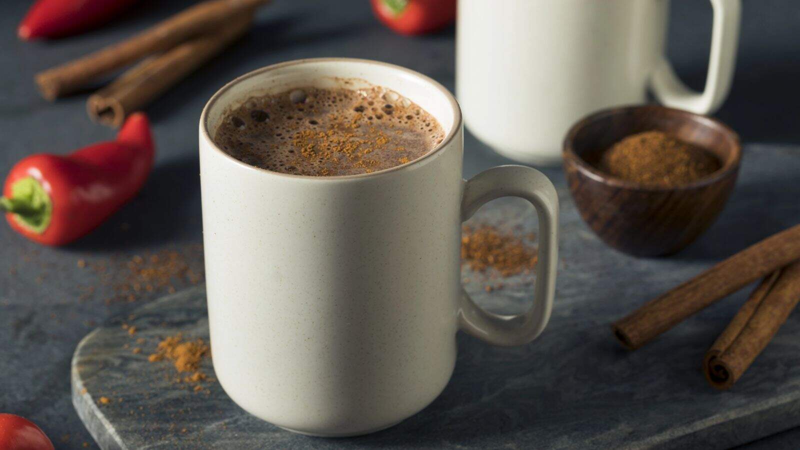 A white mug filled with hot chocolate is placed on a gray surface, surrounded by cinnamon sticks and red chili peppers. A small wooden bowl containing powdered spice is nearby. Another white mug is in the background.
