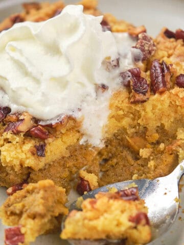 A slice of pumpkin dump cake topped with whipped cream sits on a beige plate. The cake is sprinkled with pecans, and a spoon is placed on the plate, having cut into the cake. The background includes a textured cloth and faint view of a cooling rack.