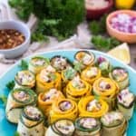 A plate of zucchini and squash rolls filled with a mixture of vegetables and herbs, arranged in a circular pattern on a turquoise dish. Surrounding the plate are small bowls containing ingredients like chopped onions, spices, and even a creamy dessert dip for an unexpected twist.