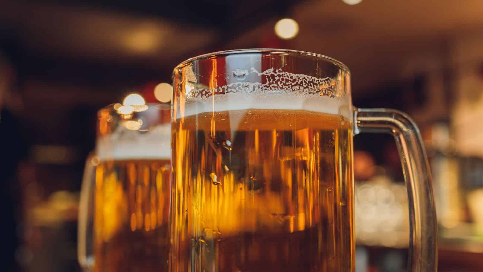 Close-up of frosty beer mugs filled with amber liquid, with light glistening on the surface. The background is softly blurred, featuring warm, ambient lighting. The focus is on the refreshing, chilled beverages.