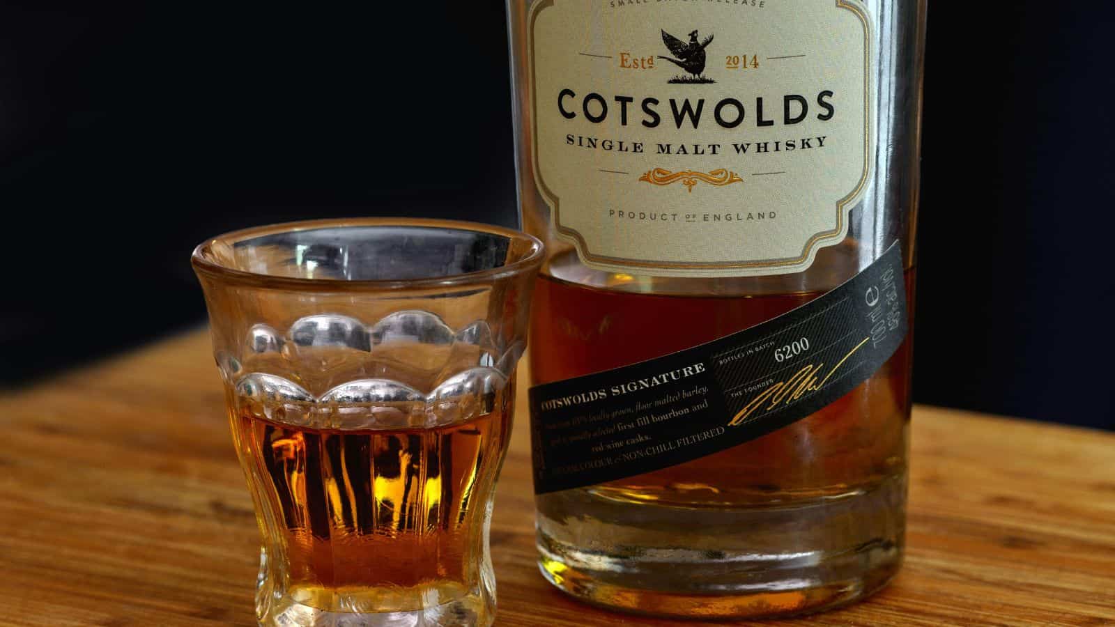 A glass of amber-colored single malt whisky sits on a wooden surface next to a bottle labeled "Cotswolds Single Malt Whisky, Product of England.