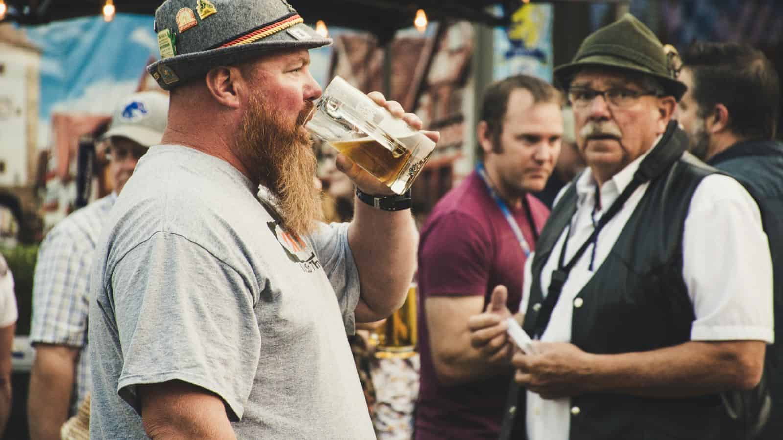 A man with a beard and gray T-shirt drinks from a glass of beer, wearing a hat with pins. He stands among a crowd at an outdoor event. Another man with glasses and a hat is in the background, talking to a group of people.