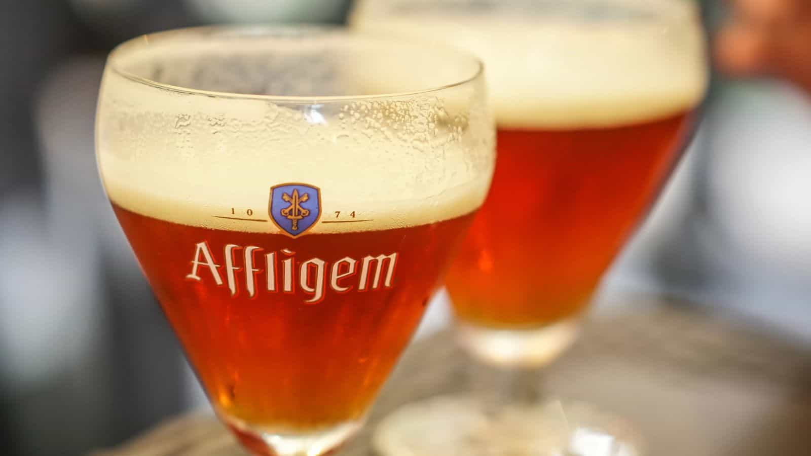Two glasses of amber-colored beer with frothy heads are placed on a table. The glasses have "Affligem" written on them with a logo. The background is blurred, focusing on the drinks.