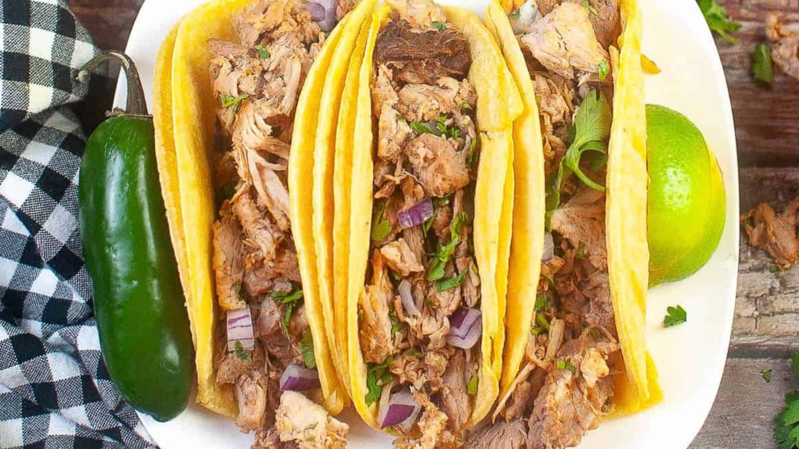 Low angle shot of 3 carnitas tacos on a white plate with lime wedges.