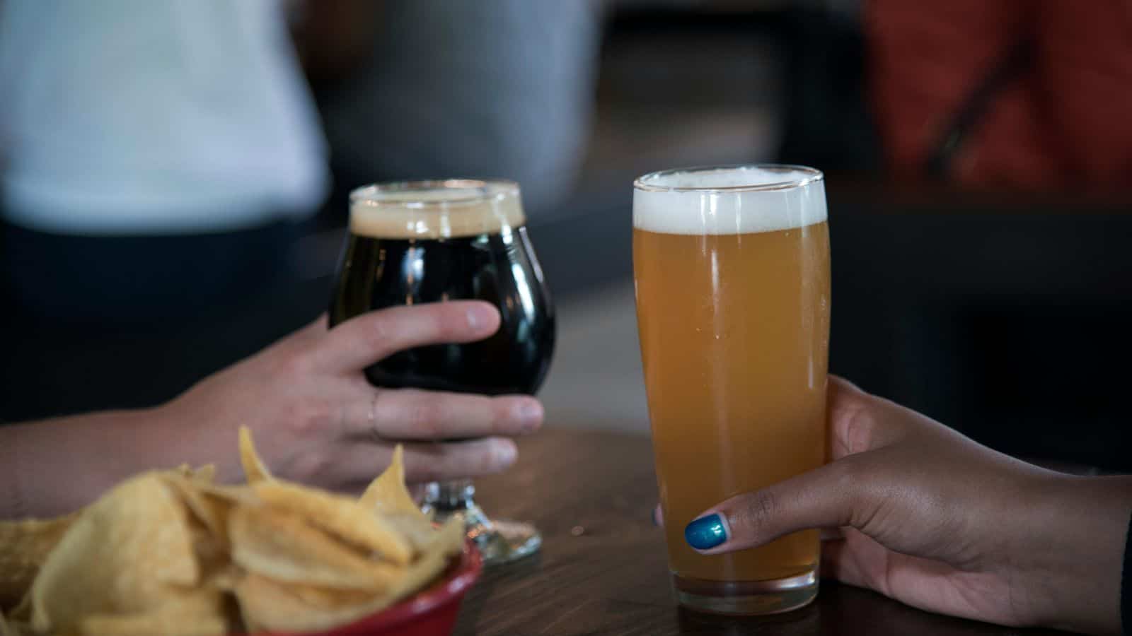 Two hands holding glasses in a pub setting. One glass contains a dark beer, while the other holds a light, amber-colored beer. A bowl of tortilla chips is visible in the foreground on a wooden table.