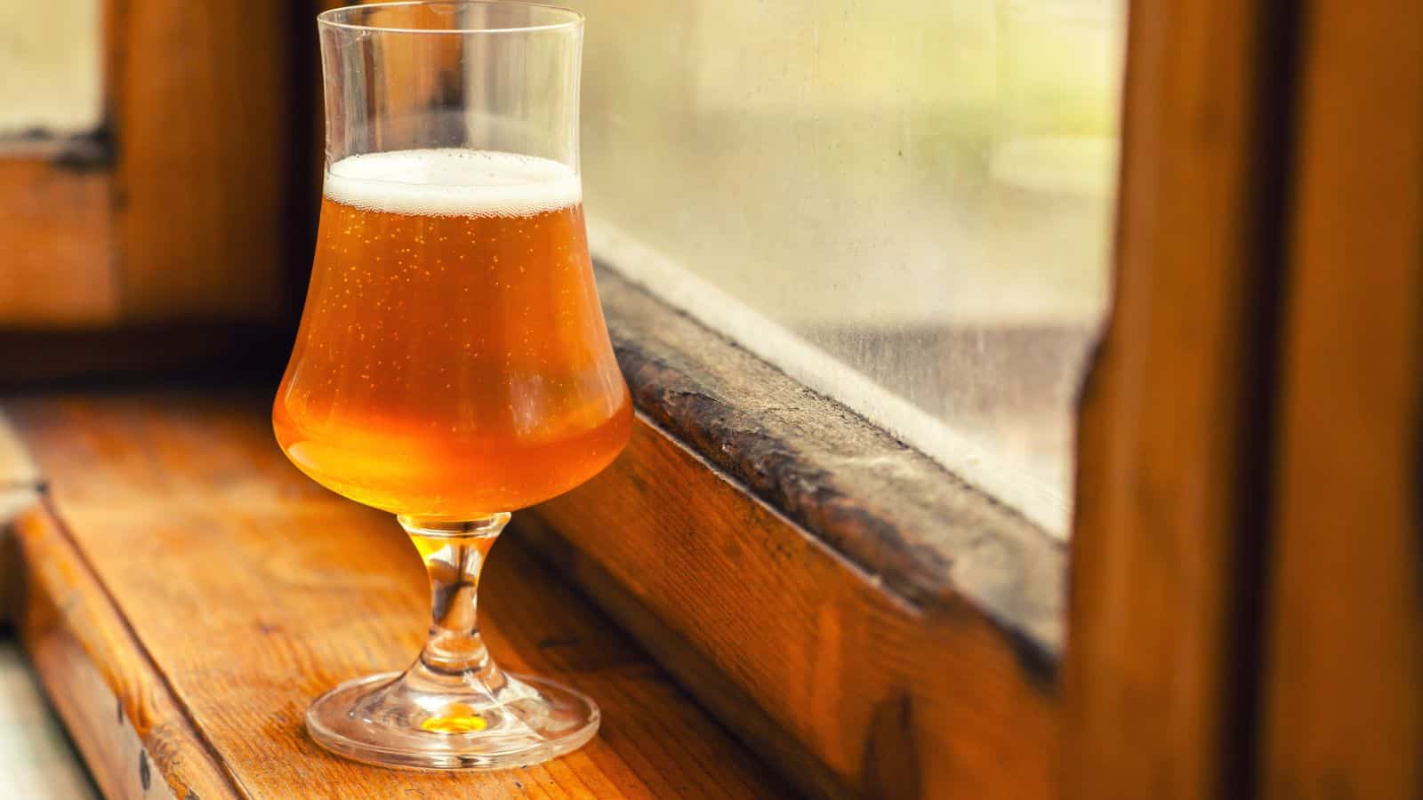 A glass of amber-colored beer with a frothy head sits on a wooden windowsill. Sunlight from the window highlights the bubbles in the drink. The glass is elegantly shaped with a thin stem.