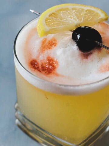 A cocktail in a glass, topped with a layer of frothy foam, garnished with a lemon wedge and a cherry on a skewer. The drink appears to be a whiskey sour, presented against a blurred blue-gray background.