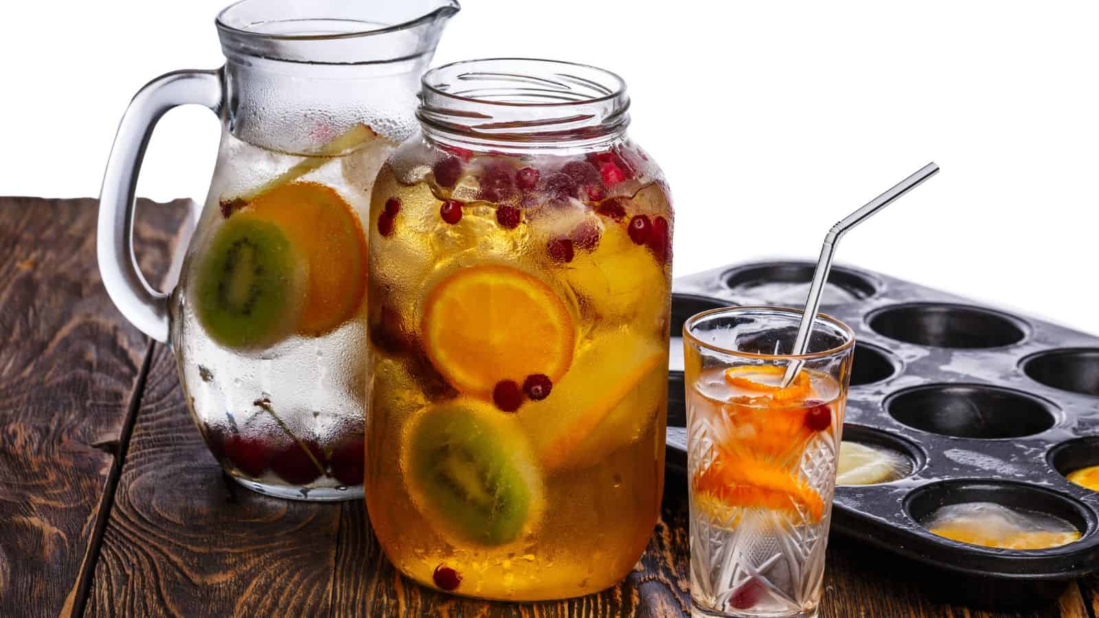 A wooden table holds a glass pitcher and a jar filled with fruit-infused water, including kiwi, orange slices, and red berries. A glass with a metal straw contains similar fruit water. A muffin tin with yellow batter is partially visible.