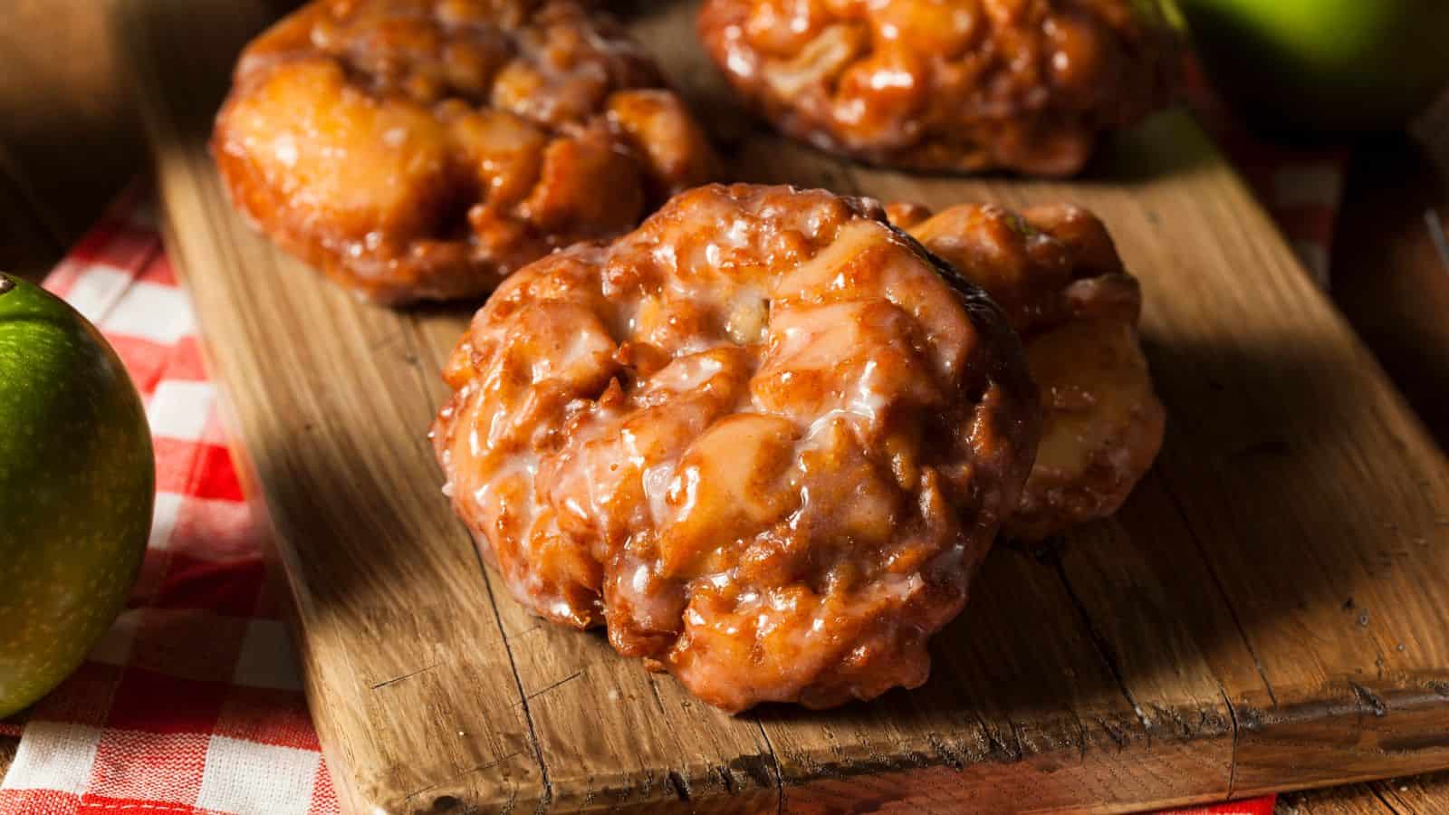 Close-up of apple fritters on a wooden board. The fritters are glazed and have a golden-brown, crispy appearance. A red and white checked cloth partially covers the surface, and a green apple is visible in the background.