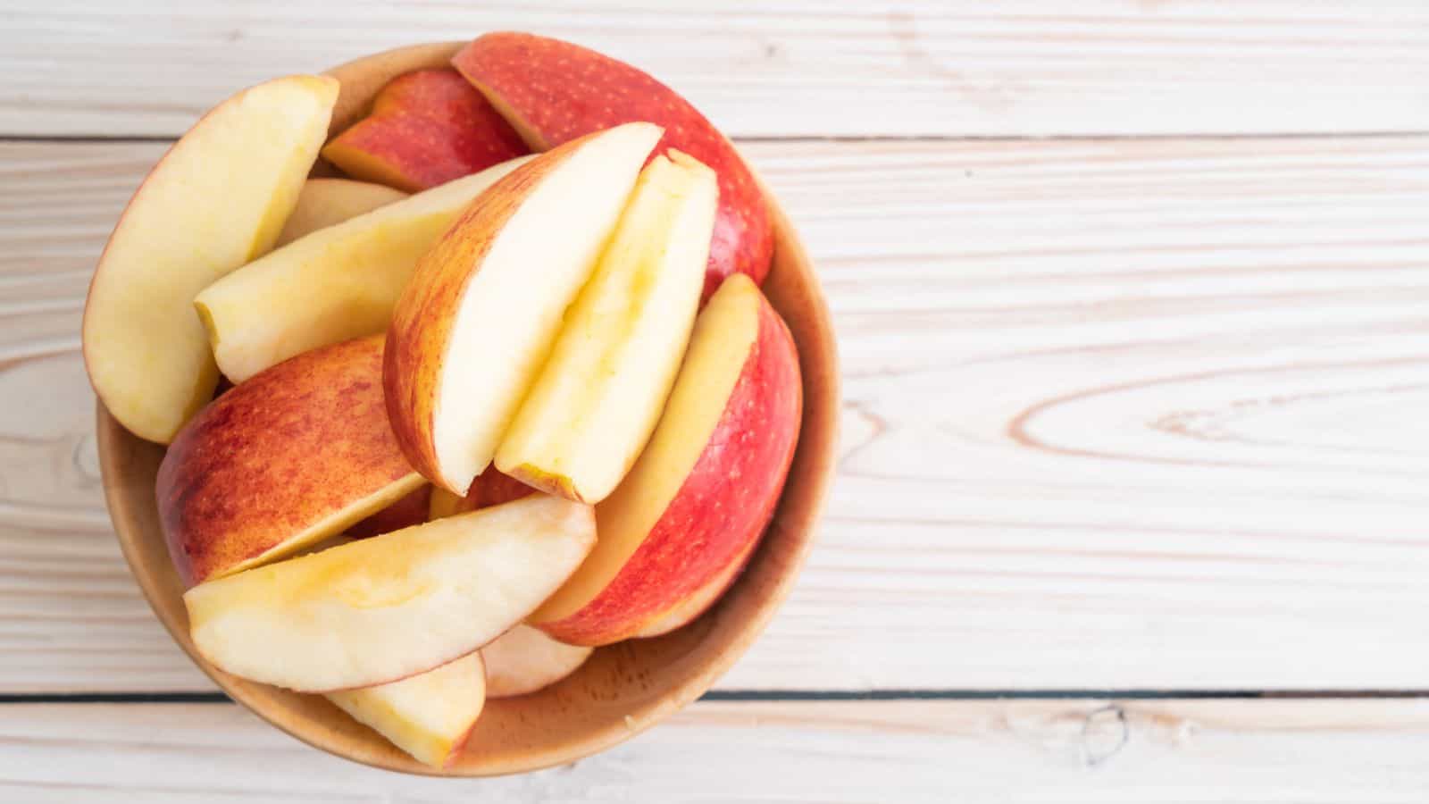 A wooden bowl filled with sliced red apples sits on a light wooden surface. The apple slices have red skin and pale yellow flesh.