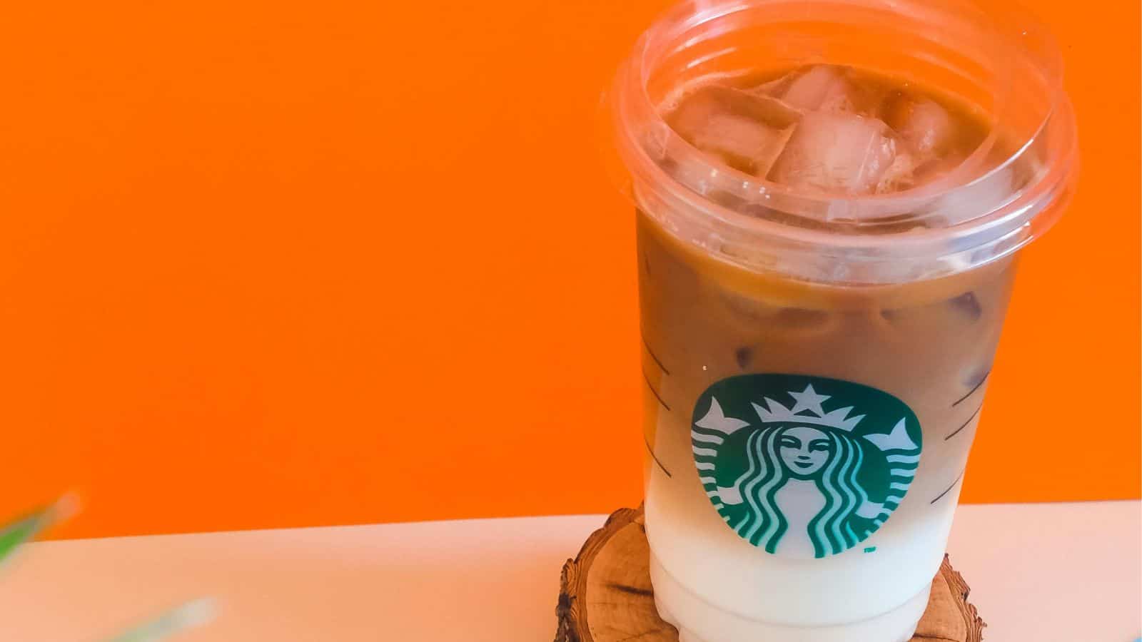 A Starbucks cold beverage in a clear cup with lid and straw is placed on a wooden coaster. The drink has a gradient, with darker coffee on top and lighter milk at the bottom. The background is bright orange.