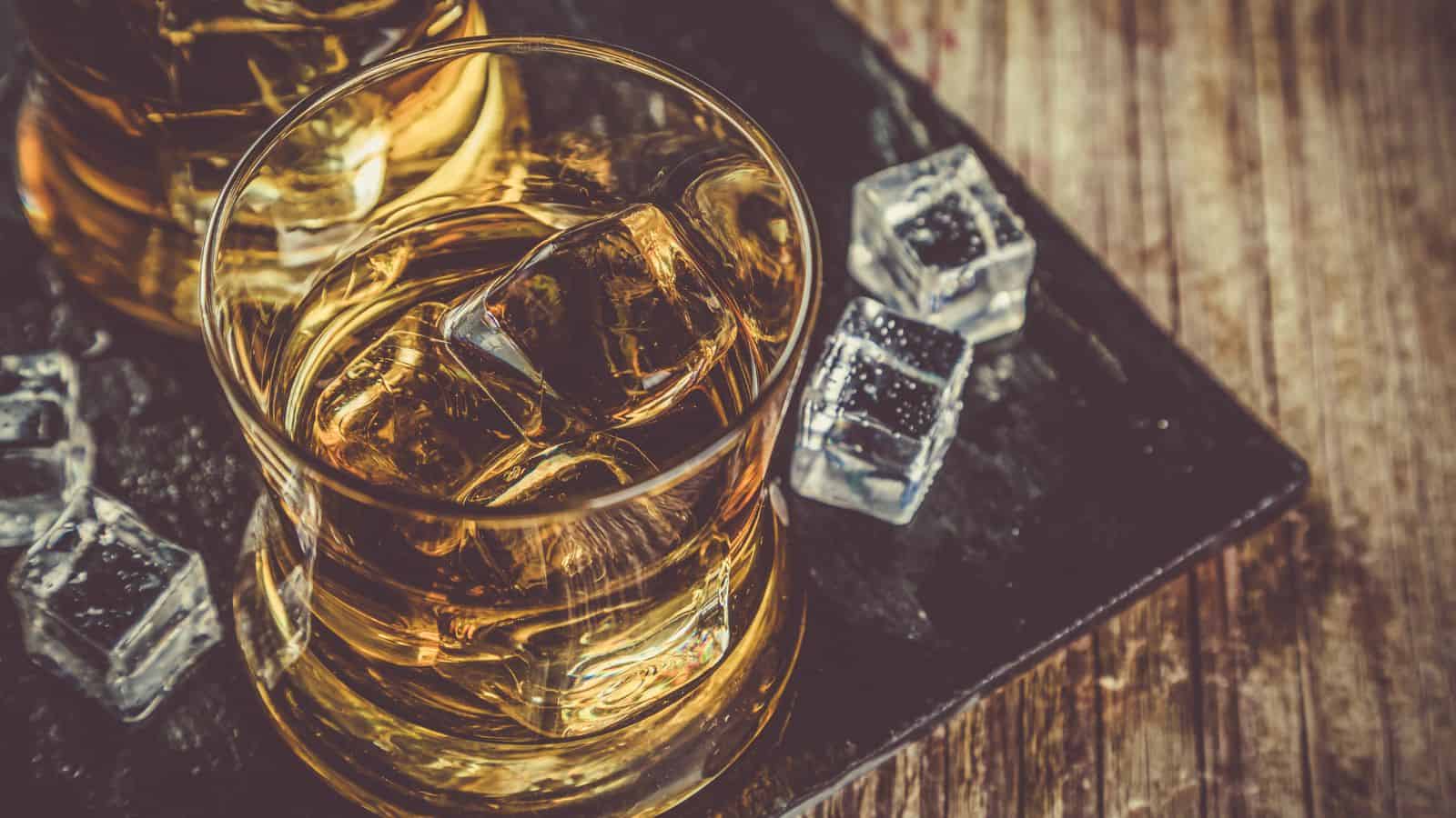 A glass of whiskey with ice cubes sits on a dark tray next to a few loose ice cubes, placed on a wooden surface. Another partially visible glass is in the upper left corner.