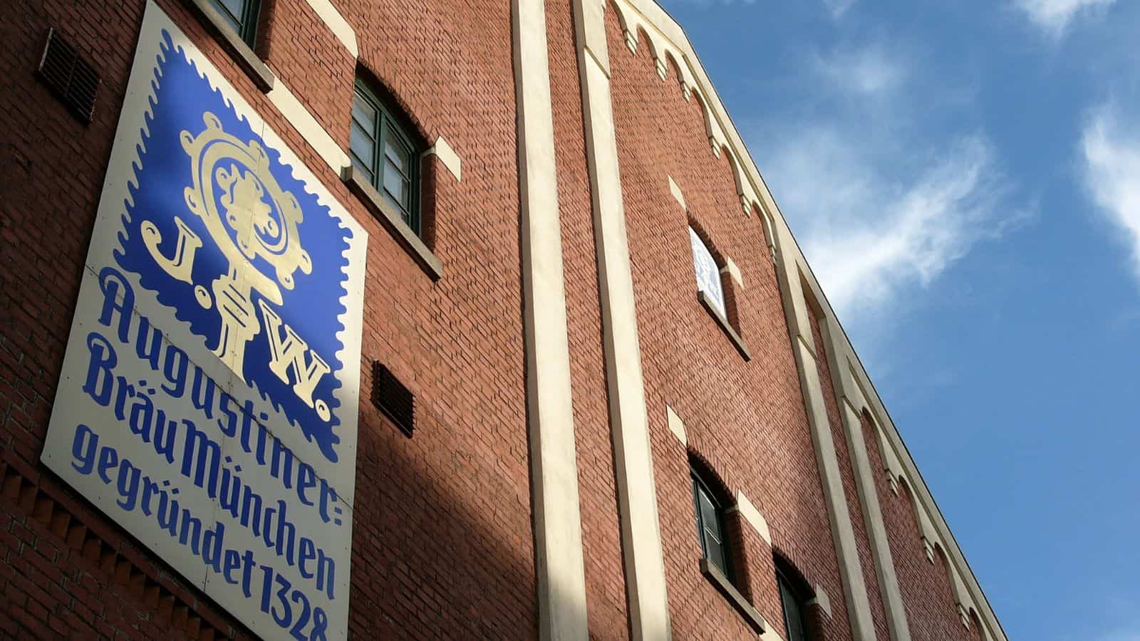 Close-up of a red brick building with a large blue and white sign displaying the logo and text "Augustiner Br&auml;u M&uuml;nchen, gegr&uuml;ndet 1328" against the facade. The sky is clear and blue in the background.