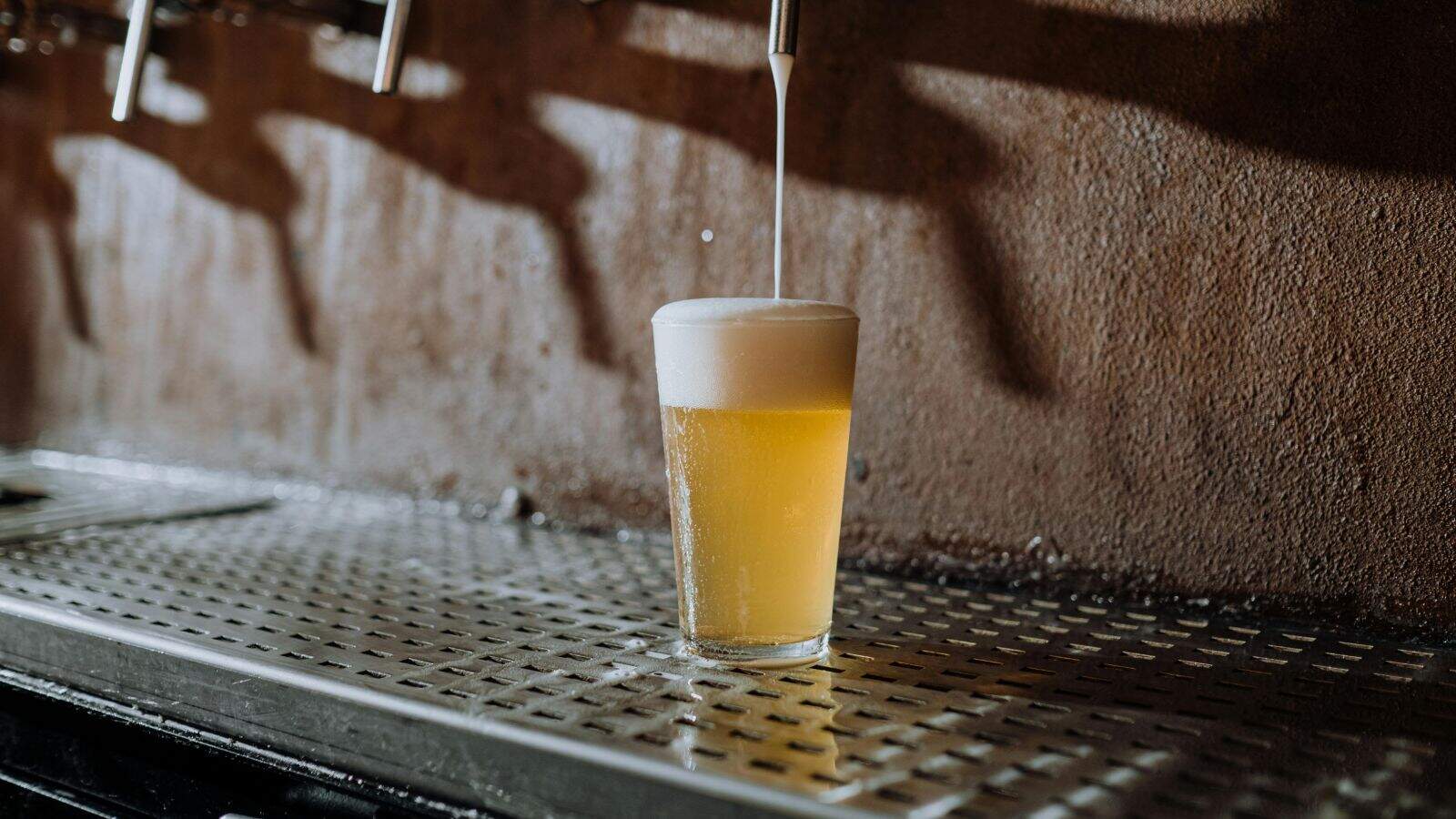 A glass of beer is being poured from a tap, with foam rising to the top of the glass. The glass sits on a perforated metal drip tray, and the background shows a brown textured wall.