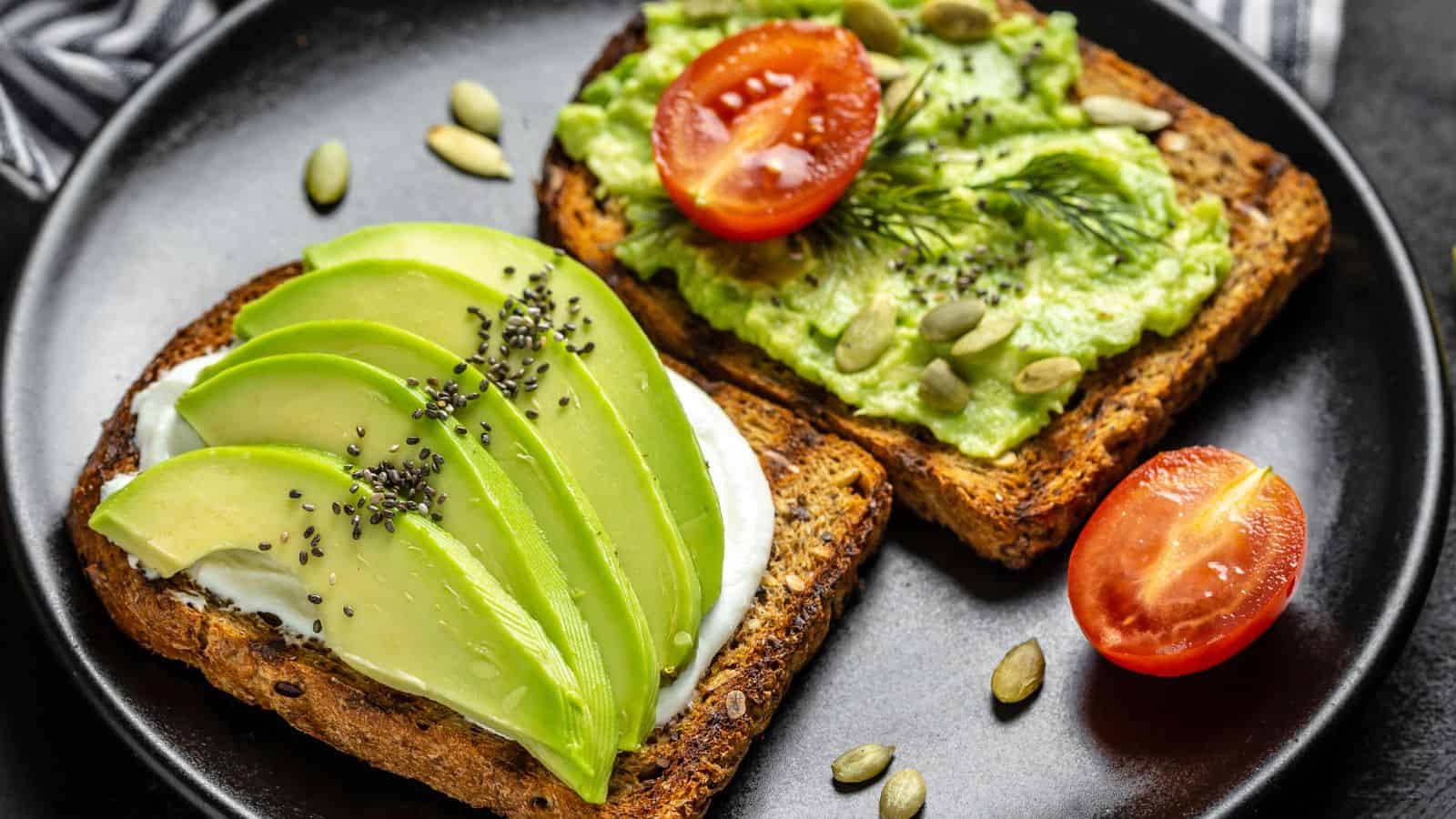Two slices of whole grain toast on a black plate; one topped with sliced avocado, a white spread, and chia seeds, the other with mashed avocado, tomato, dill, and pumpkin seeds. A halved cherry tomato lies beside them.