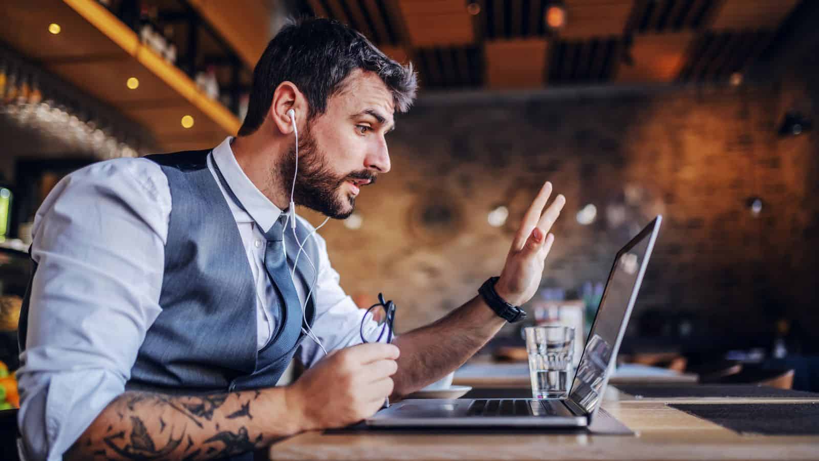 A bearded man in a vest and tie sits at a table in a caf&eacute;, engaged in a video call on a laptop. He gestures with one hand while holding glasses in the other. Earbuds connect him to the device. A glass of water is on the table.