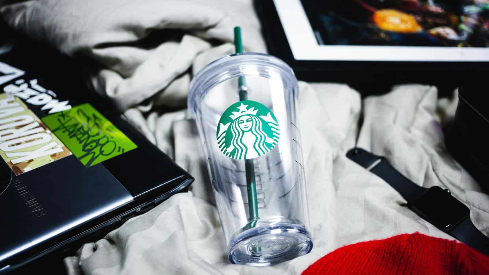 A clear Starbucks tumbler with a green straw lies on crumpled light-colored fabric. Nearby are a closed laptop, a tablet, and a red item. The tumbler features the iconic Starbucks logo.