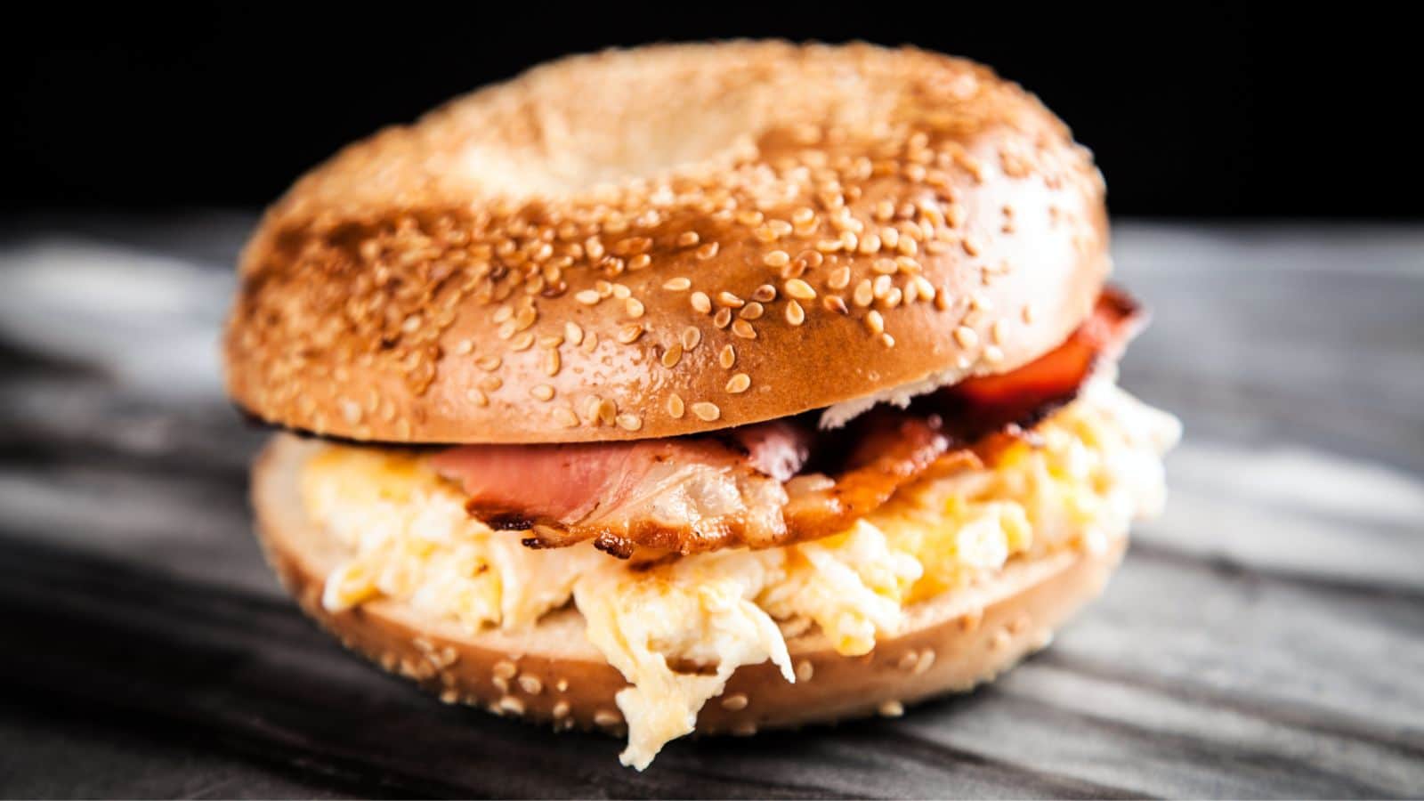 A sesame seed bagel sandwich filled with scrambled eggs and crispy bacon sits on a gray marble surface.
