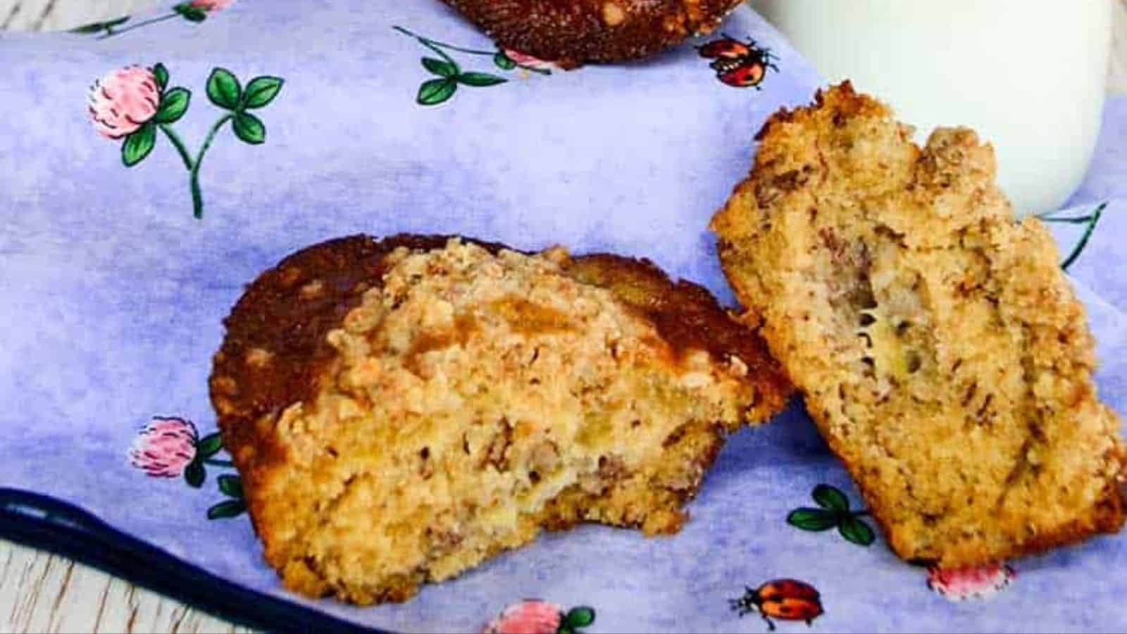 Two pieces of banana nut muffin with crumb topping are placed on a light blue cloth decorated with flowers and ladybugs. A glass of milk is partially visible in the background.