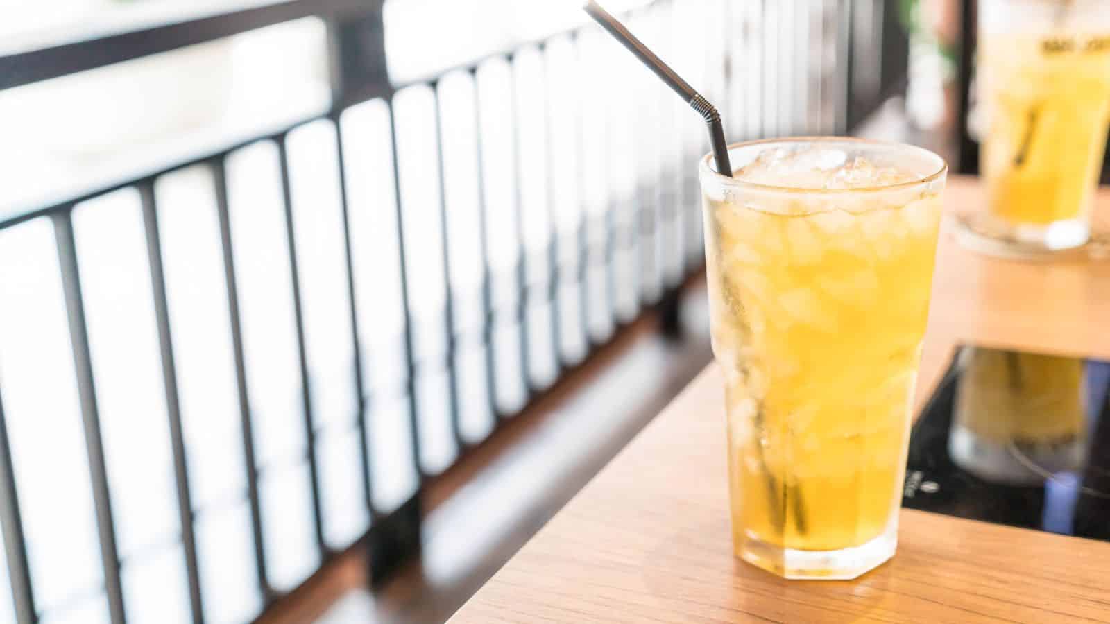 A tall glass filled with iced yellow liquid, possibly a juice or tea, sits on a wooden table. A black straw is placed in the glass. Another similar drink is in the background. Sunlight filters through a railing beside the table.